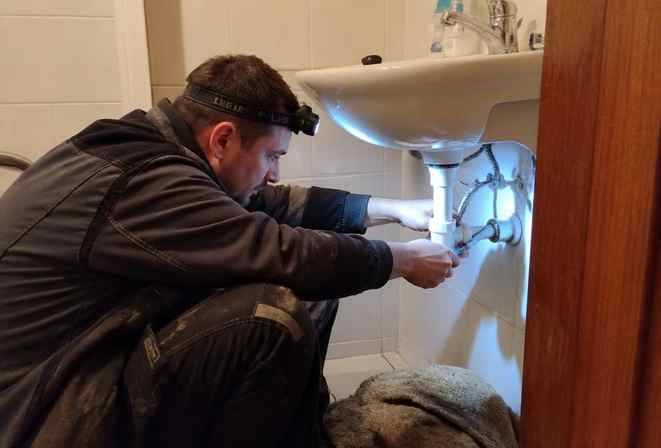 Instalatér s čelovkou opravuje sifon pod umyvadlem v koupelně
